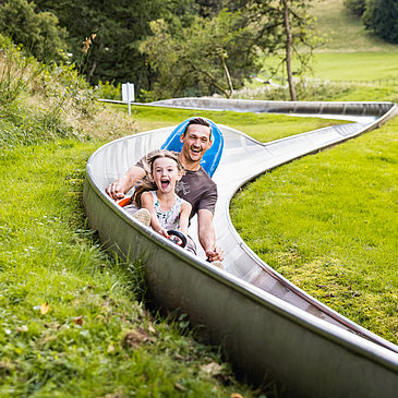 Das Bild zeigt ein Mann und ein Mädchen, die eine Sommerrodelbahn fahren.